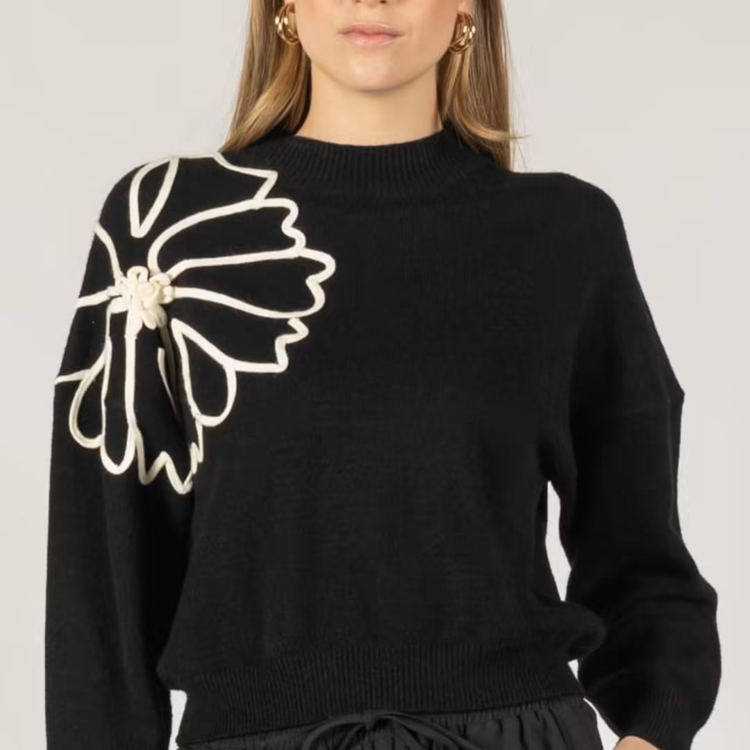Wearing a black knit sweater with a large white floral applique on the left shoulder.